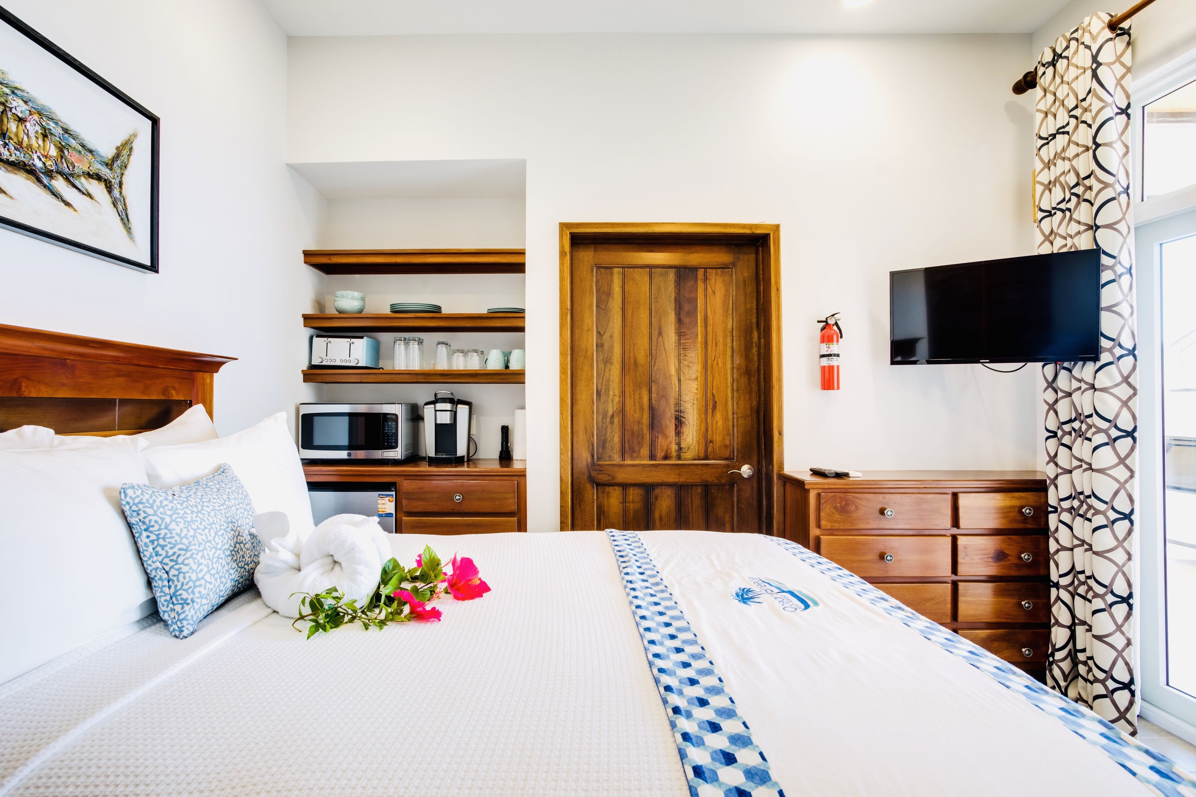 A neatly arranged hotel room with a bed, a television, a wooden door, and a shelf with kitchen items.