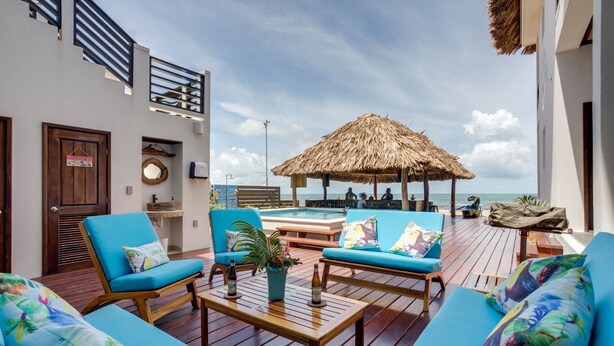 A sunlit outdoor lounge area with wooden flooring, blue cushioned seating, a thatched umbrella, and a view of the ocean. - Ocean Breeze (Placencia)