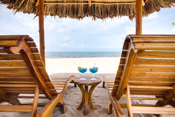 A wooden beach chair and umbrella setup with a table and two drinks in the sand, overlooking the ocean. - Ocean Breeze (Placencia)