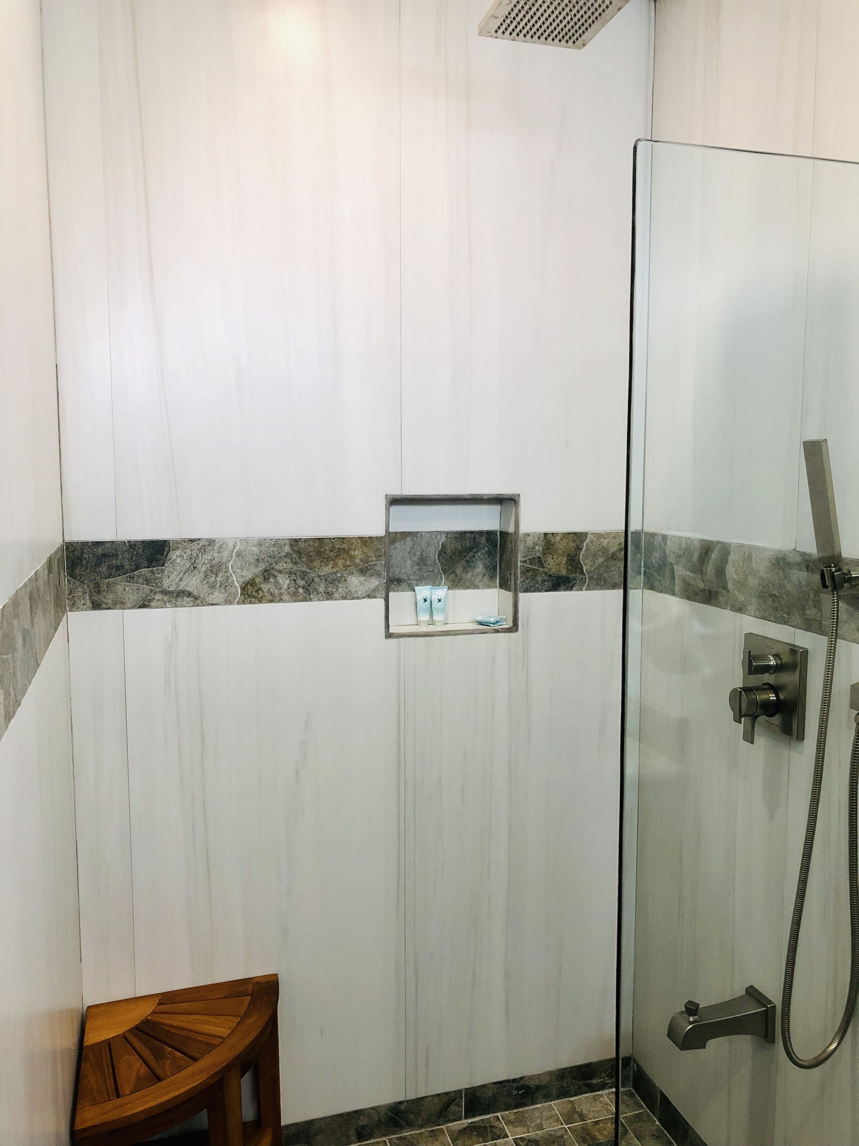 A bathroom with a glass shower enclosure, a wooden stool, and a small mirror with toiletries.