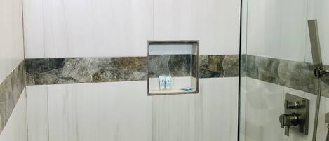A bathroom with a glass shower enclosure, a wooden stool, and a small mirror with toiletries.