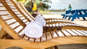 A wooden lounge chair with a rolled-up towel on it, a blue table, and a colorful bird-shaped chair in the background. - Ocean Breeze (Placencia)