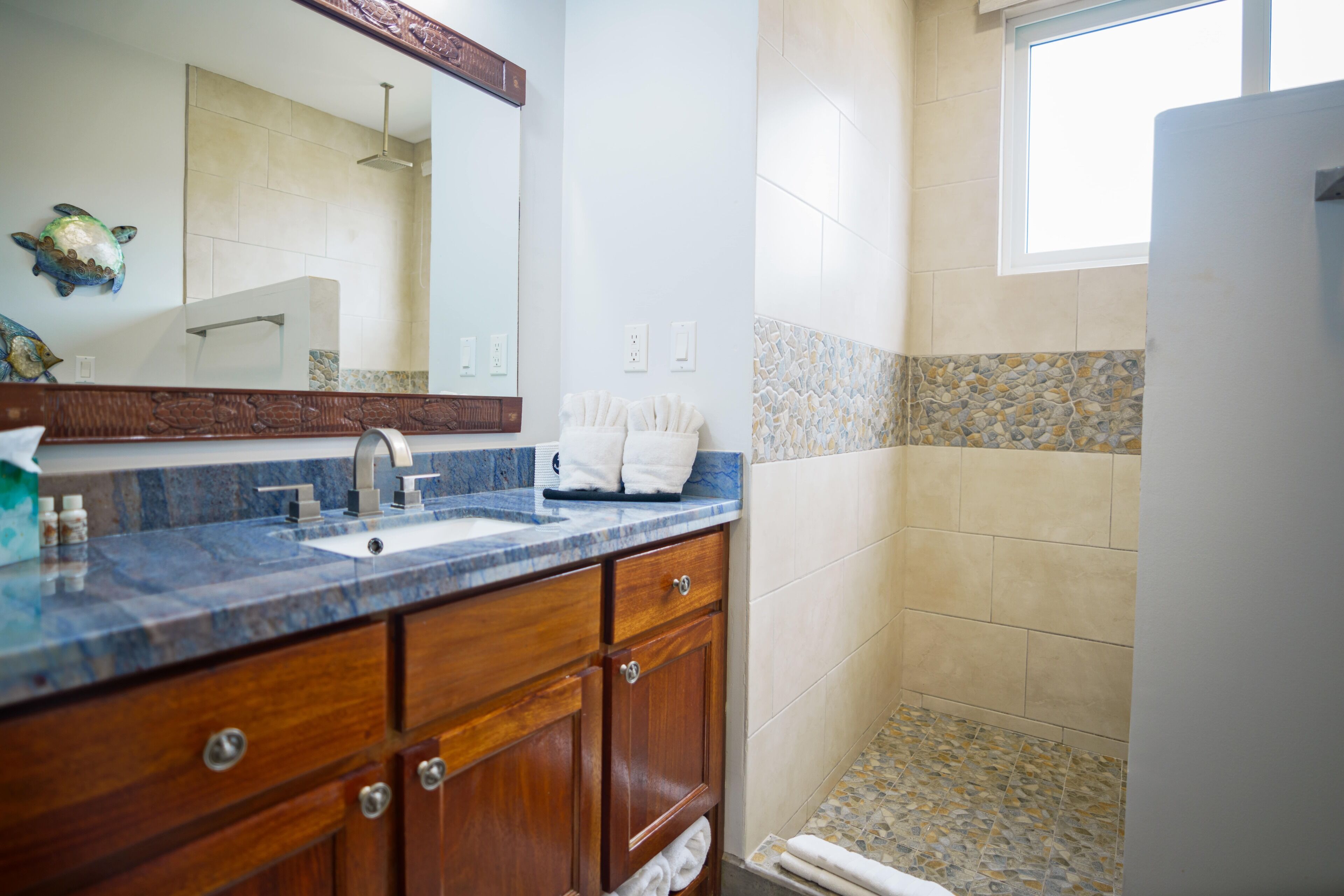 A bathroom with a marble countertop, wooden cabinets, a large mirror, and a shower area with mosaic tiles.
