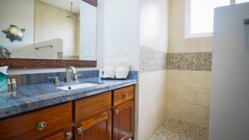 A bathroom with a marble countertop, wooden cabinets, a large mirror, and a shower area with mosaic tiles.