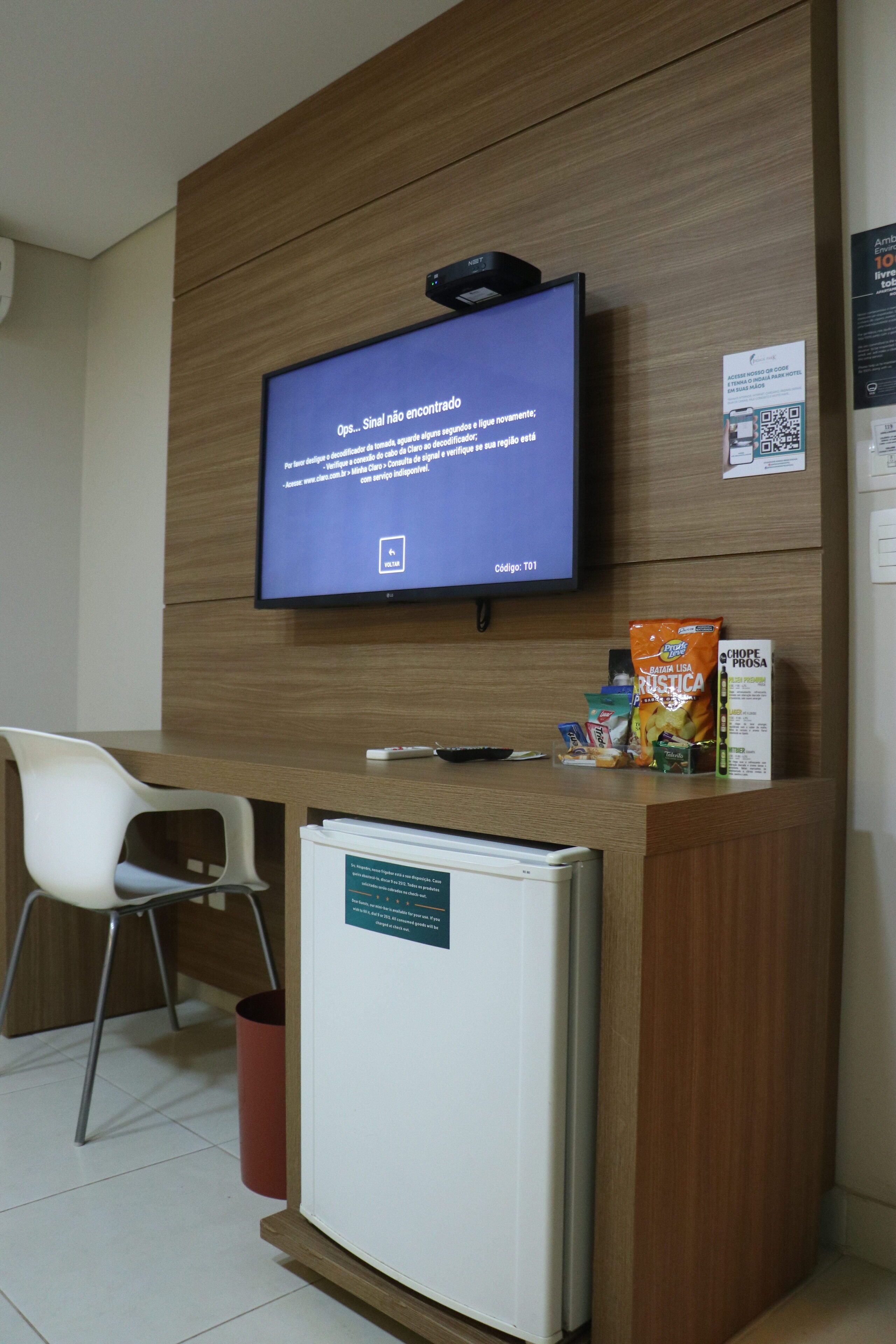 Un mostrador de recepción con un televisor de pantalla plana que muestra un mensaje, un refrigerador pequeño, una silla y algunos aperitivos.