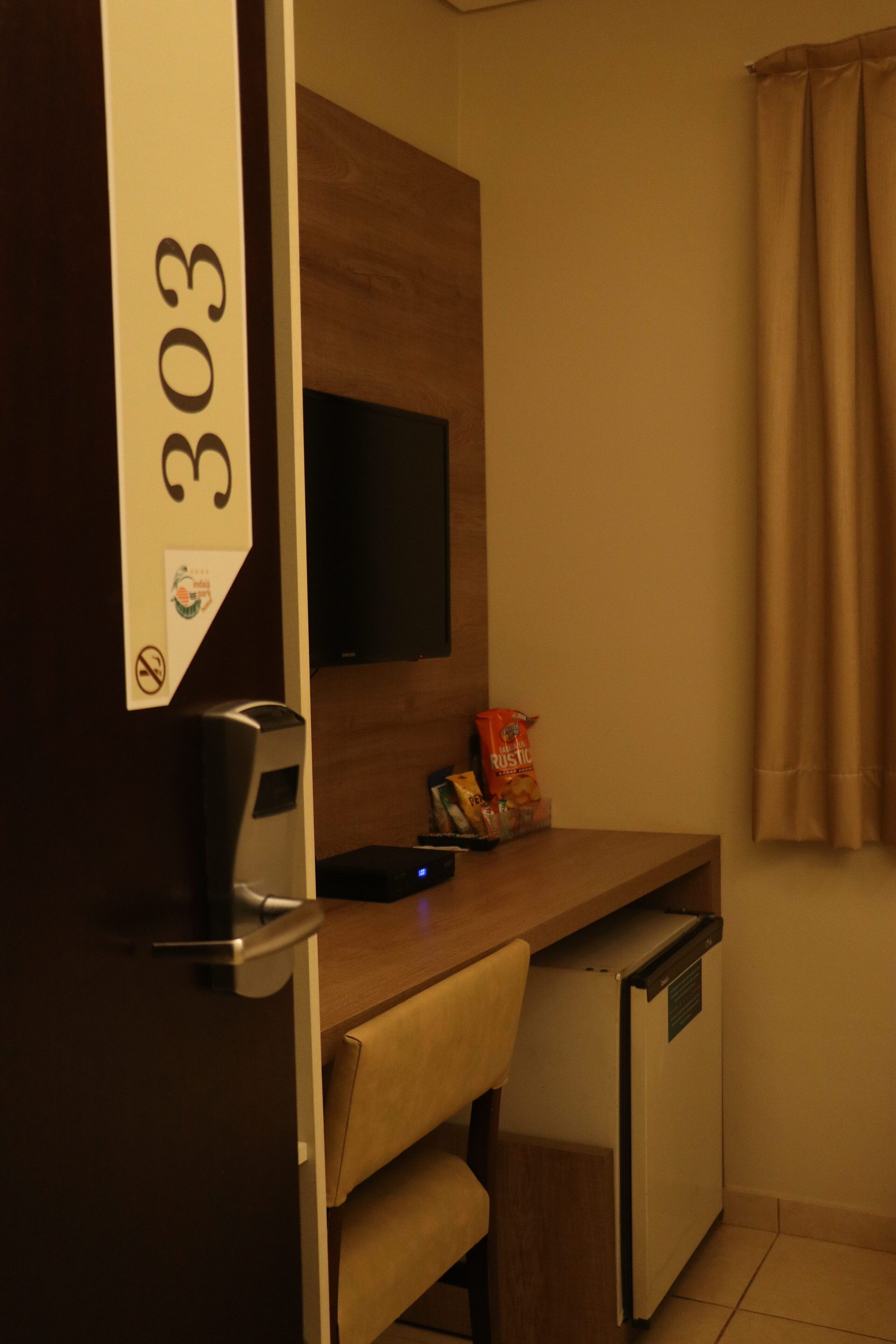 Una habitación compacta con un escritorio de madera, un refrigerador pequeño, un televisor montado en la pared y una cortina.
