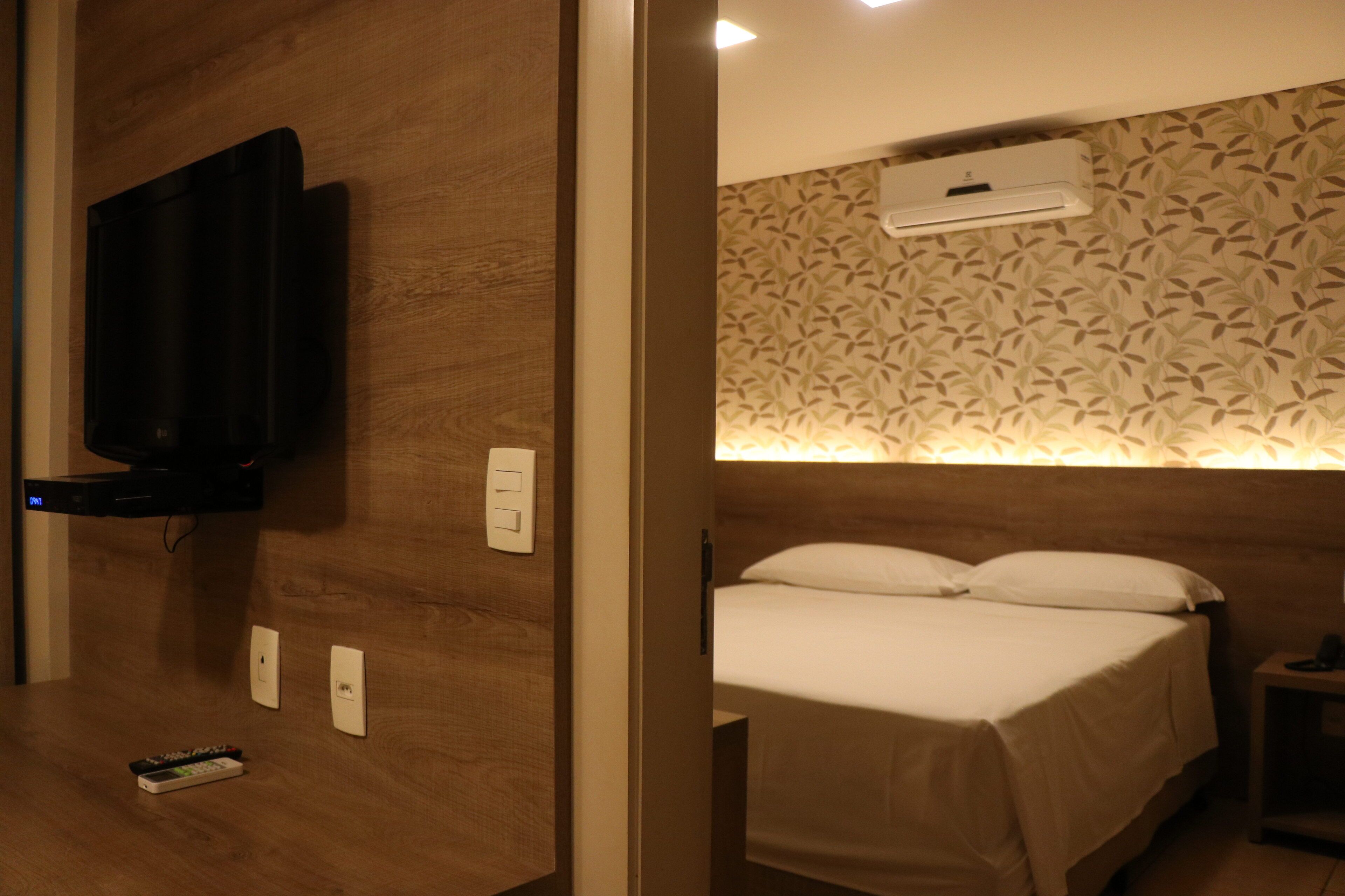 A hotel room with a bed, a TV mounted on the wall, a bedside table, and a wall-mounted air conditioner.