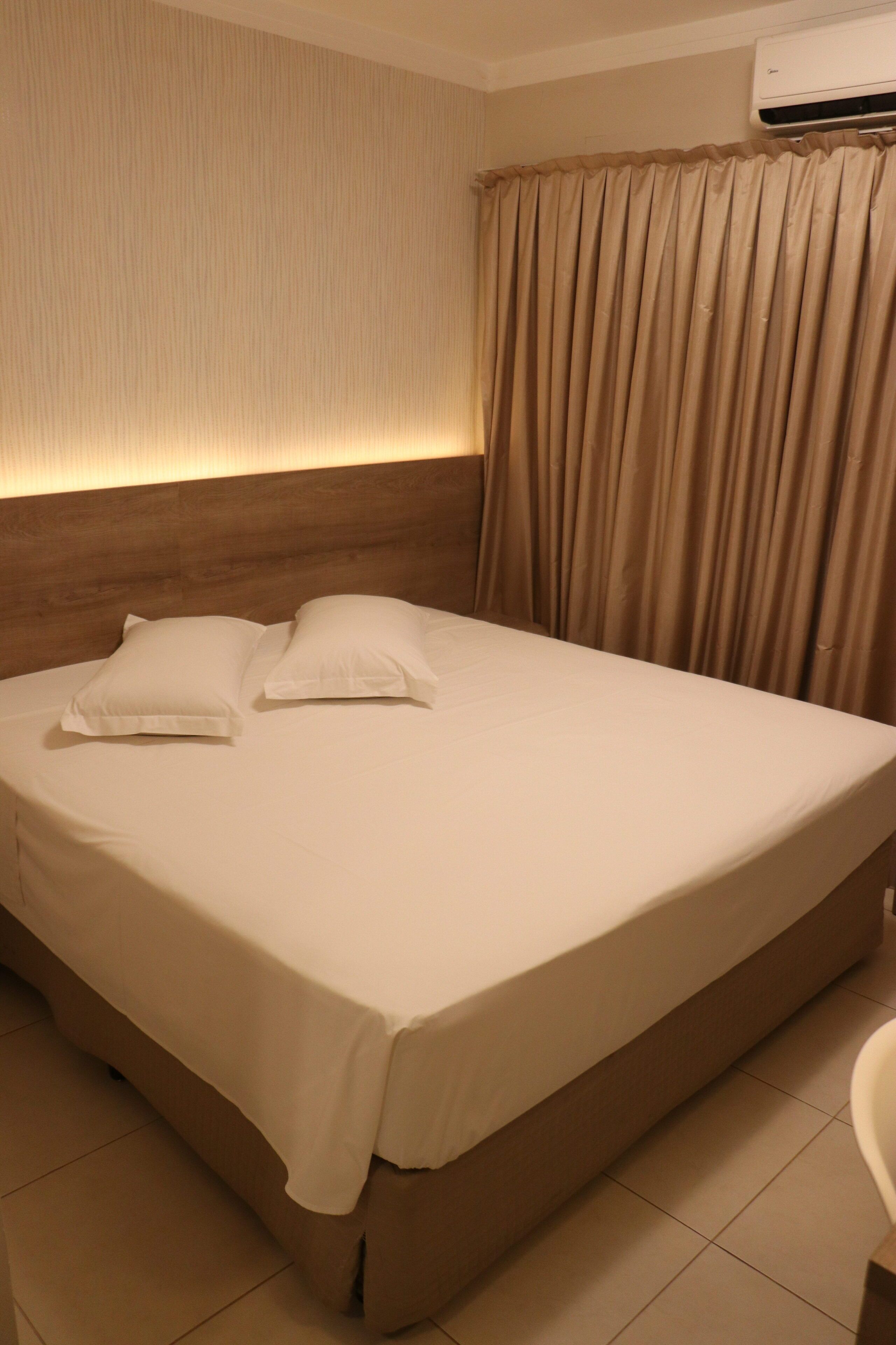 Una cama bien hecha con sábanas blancas y dos almohadas, cabecera de madera, cortinas color beige y suelo de baldosas.