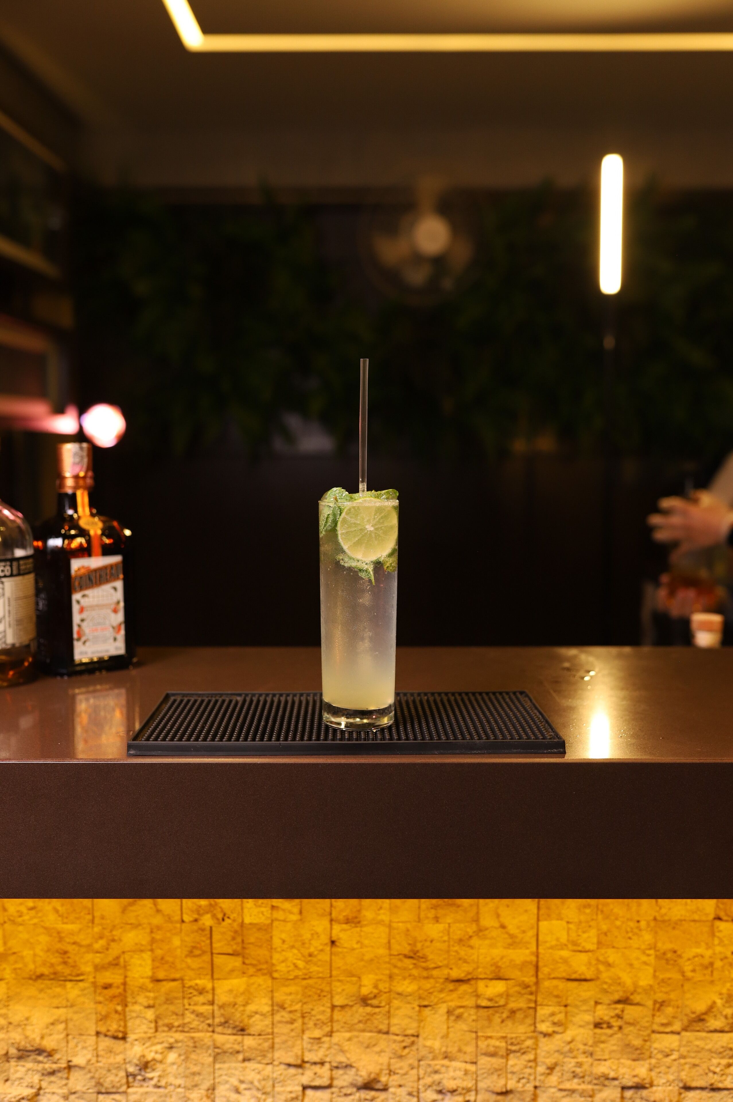 A bar counter with a tall glass of a clear beverage garnished with lime and mint, a bottle of liquor, and a bottle of water.