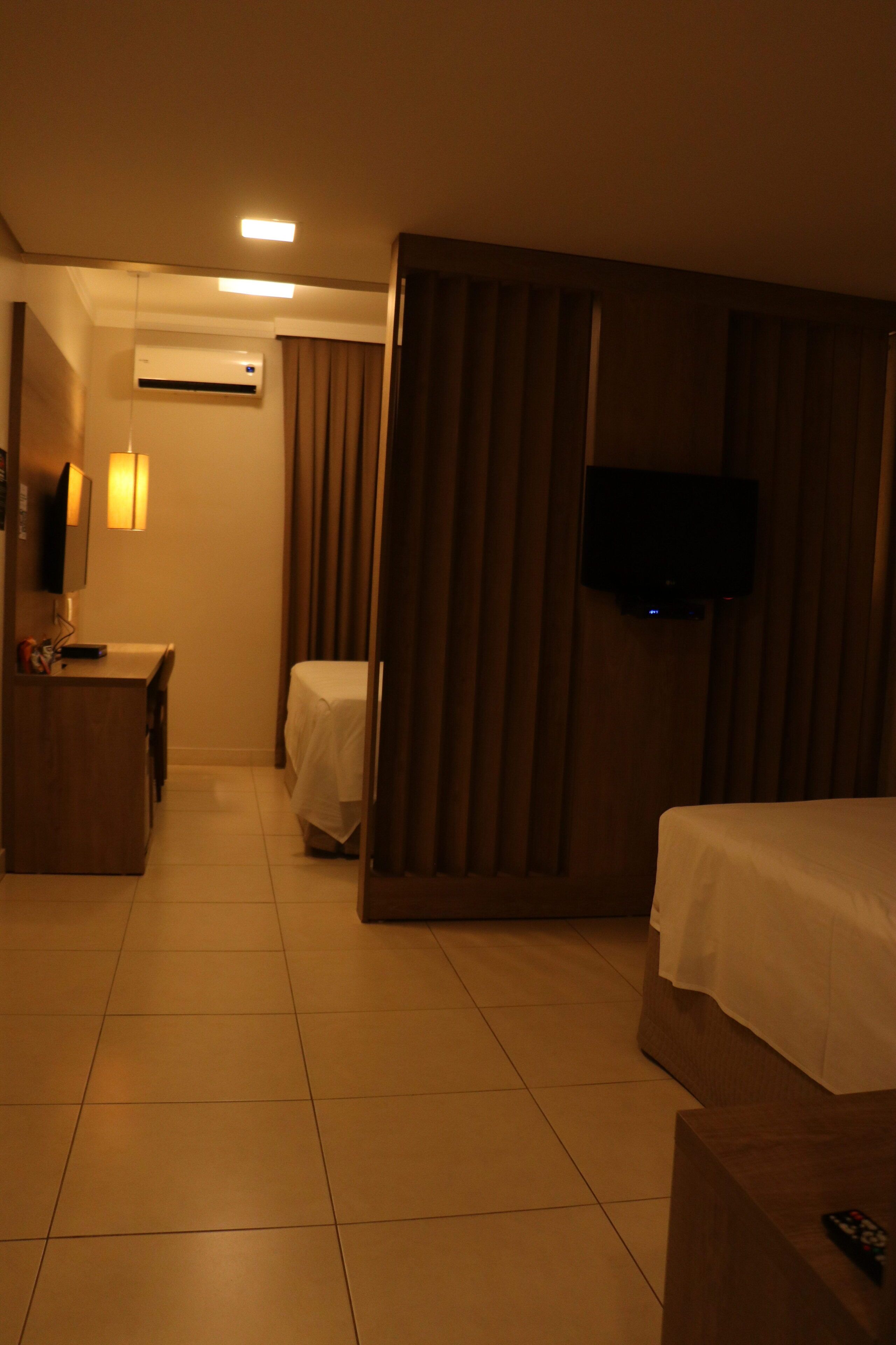 Quarto de hotel com cama, escrivaninha, cadeira, televisão e ar condicionado.