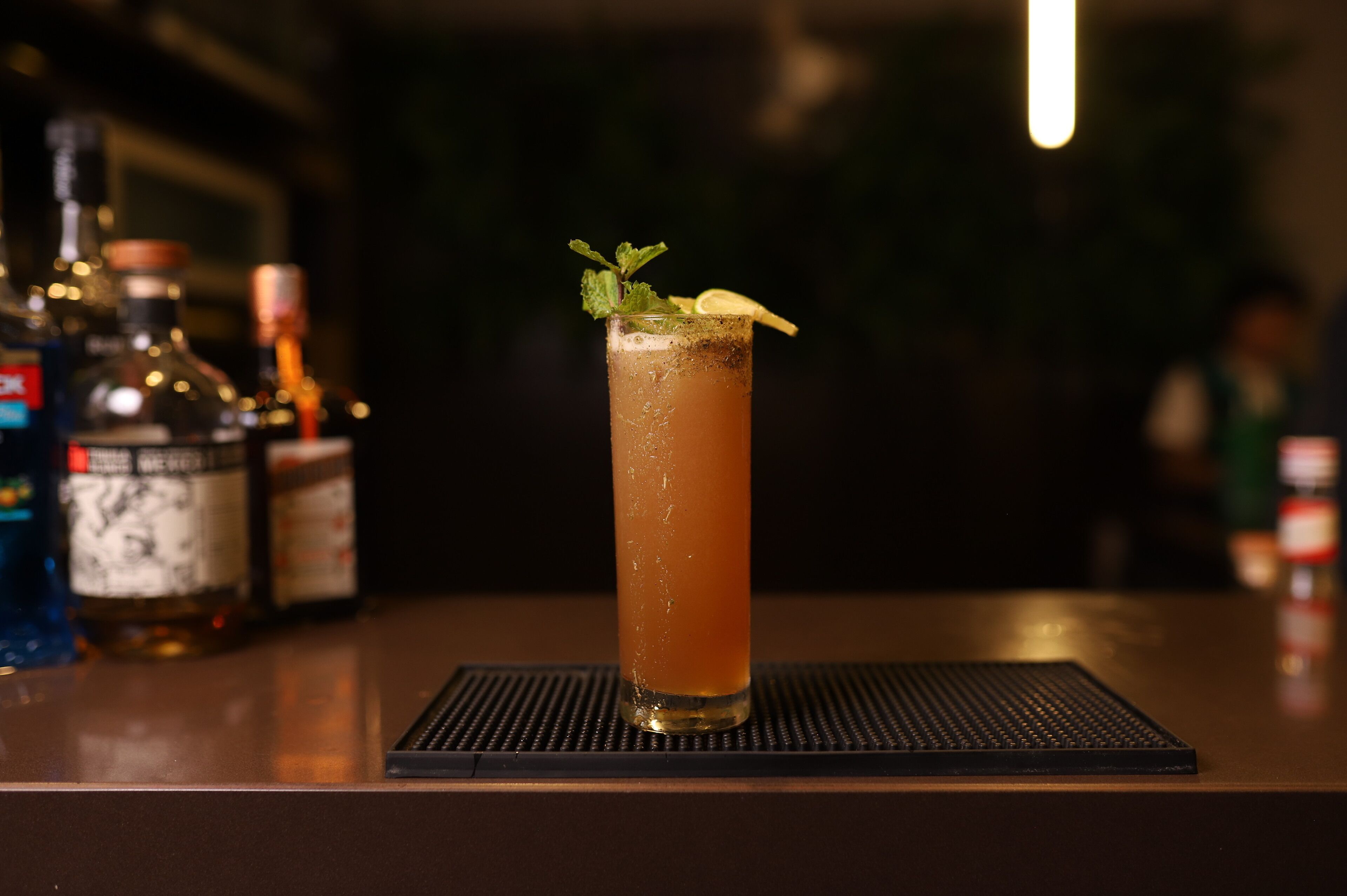 A tall glass of amber-colored drink with a mint garnish on a bar counter, surrounded by bottles of liquor.
