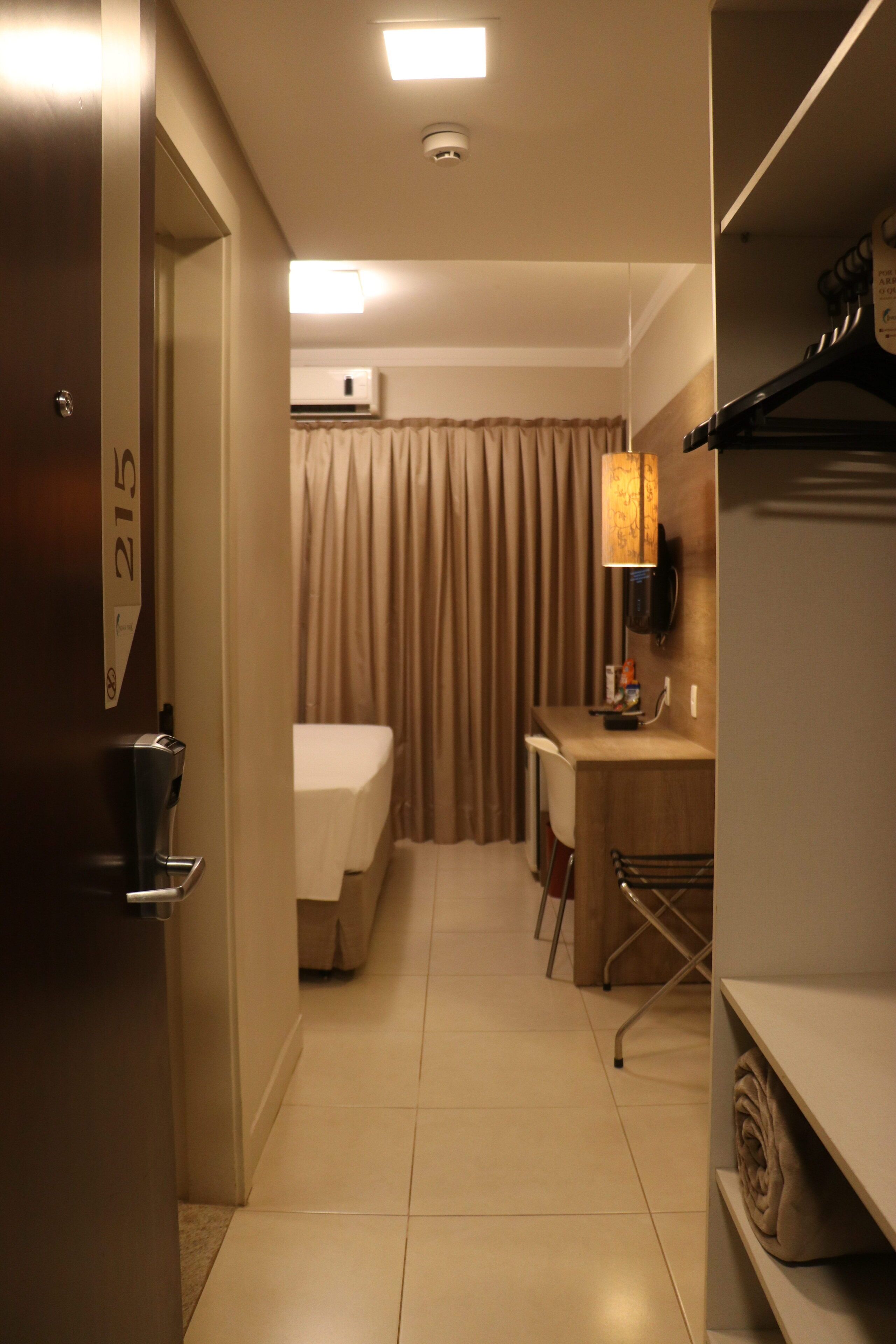 Quarto de hotel com cama, escrivaninha, cadeira, luminária e armário.