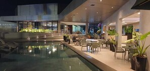 Een moderne hotel lobby met een zwembad, buitenzitplaatsen en een trap naar een hoger niveau.