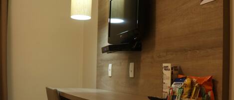 A compact hotel room with a kitchenette, a television mounted on the wall, a small refrigerator, a chair, and a hanging light fixture.