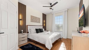 Un dormitorio con cama, mesitas de noche, ventilador de techo, televisor y ventana con persianas.