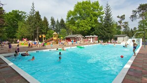 Una piscina con persone che nuotano e giocano, circondata da alberi e un'area giochi.