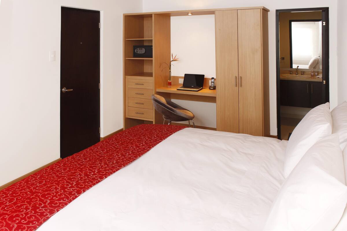 A bedroom with a bed, a desk with a chair, a wardrobe, and a bathroom with a mirror.