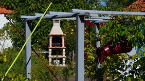 En trädgård med en grillplats i tegel under en pergola, omgiven av grönska och blommor.