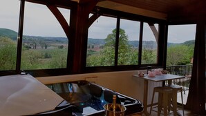 Una habitación con un ventanal grande que ofrece vistas a un paisaje verde y colinas. Hay una mesa de madera con una botella de champán y una mesa pequeña con un objeto rosa encima.