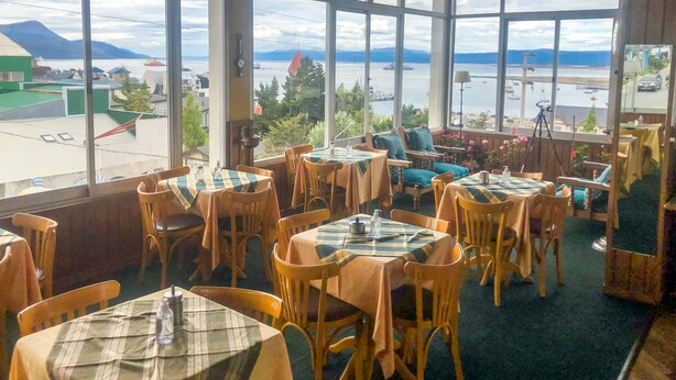 Un restaurante con amplios ventanales que ofrece vistas a un cuerpo de agua y montañas. Las mesas están cubiertas con manteles a cuadros y sillas de madera.