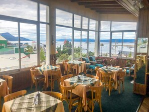 Un restaurante con amplios ventanales que ofrece vistas a un cuerpo de agua y montañas. Las mesas están cubiertas con manteles a cuadros y sillas de madera.