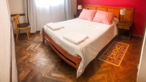 Una cama bien hecha con sábanas blancas, cabecera de madera y almohadas rosas. Hay una silla de madera y una mesita de noche con una lámpara. El piso es de madera con patrón de espiga y tiene una alfombra estampada.