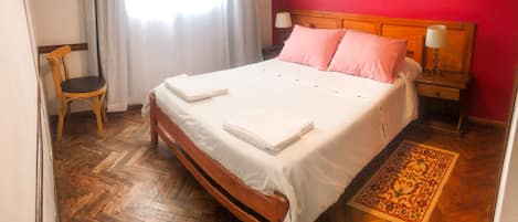 Cama bem arrumada com lençóis brancos, cabeceira de madeira e almofadas cor-de-rosa. Há uma cadeira de madeira e uma mesa de cabeceira com abajur. O quarto tem piso de madeira em padrão herringbone e um tapete estampado.
