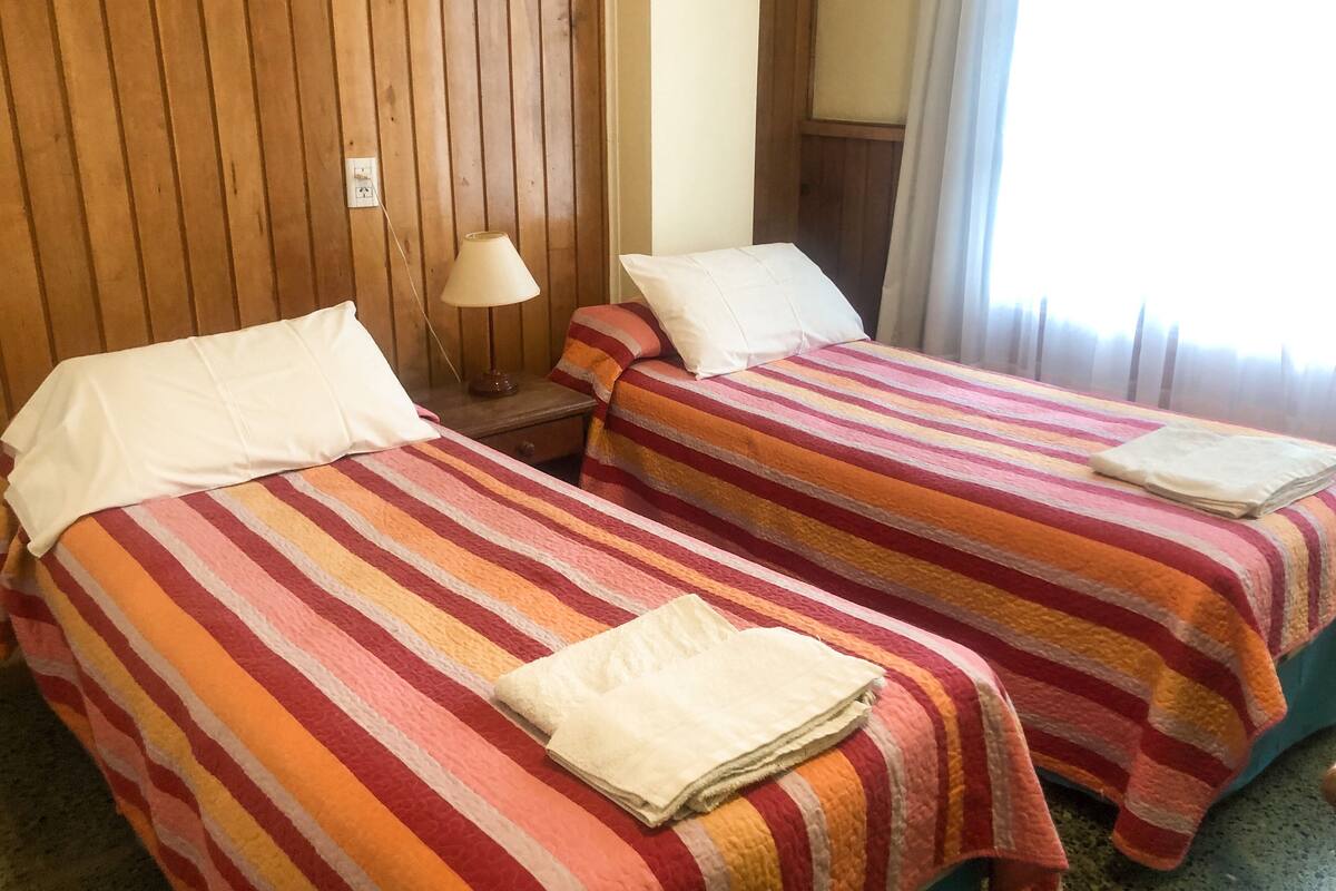 Habitación de hotel con dos camas, cada una con ropa de cama y almohadas a rayas. Hay una mesita de noche con una lámpara entre las camas.