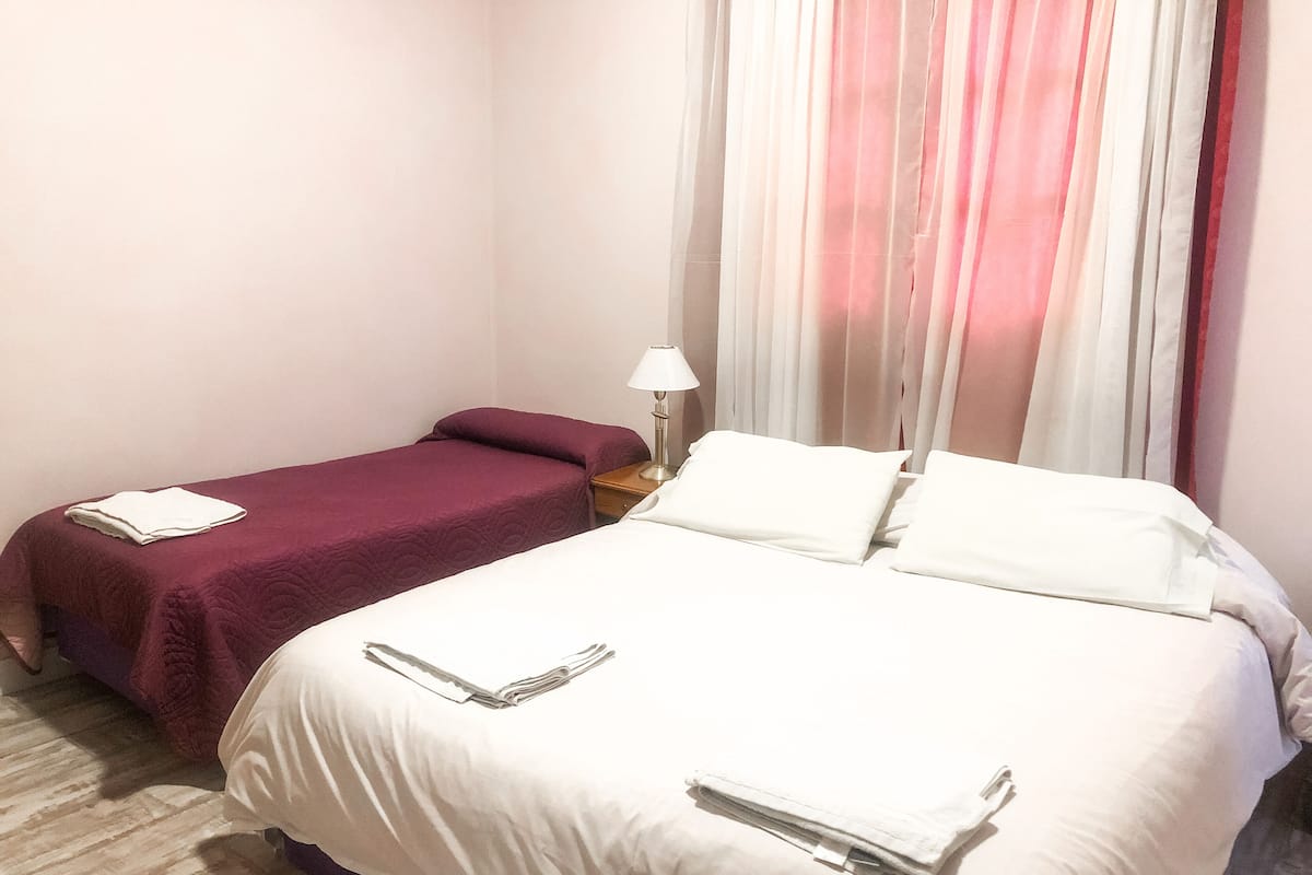 Habitación de hotel con dos camas, una con cubrecama blanco y la otra con cubrecama color burdeos. Entre las camas, un mesita de noche con una lámpara.