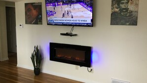 A living room with a flat-screen TV mounted on the wall, a fireplace below it, and a potted plant to the left.