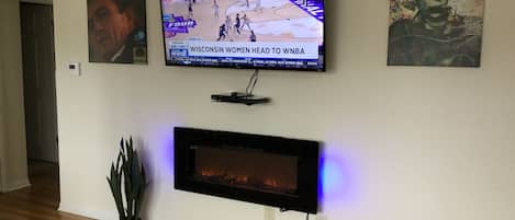 A living room with a flat-screen TV mounted on the wall, a fireplace below it, and a potted plant on the floor.
