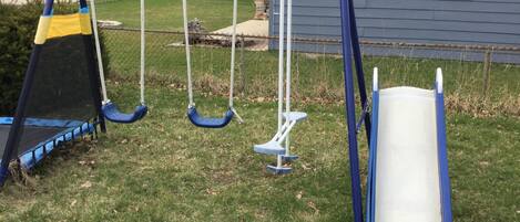 A playground with swings and a slide, set against a house and lawn.