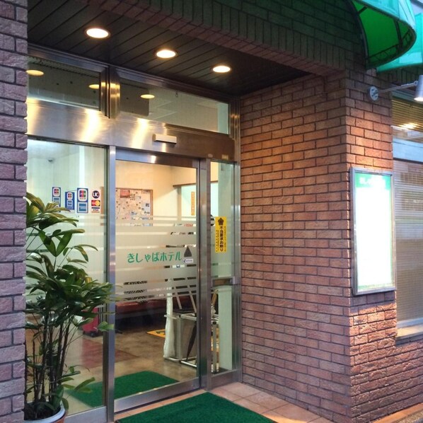 Entrada de un hotel con puerta de vidrio, pared de ladrillo y toldo verde.