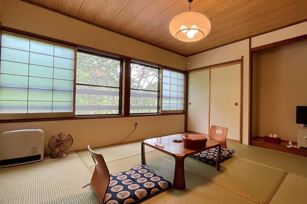 A traditional Japanese room with tatami flooring, a low table, cushions, a fan, a television, and sliding doors.