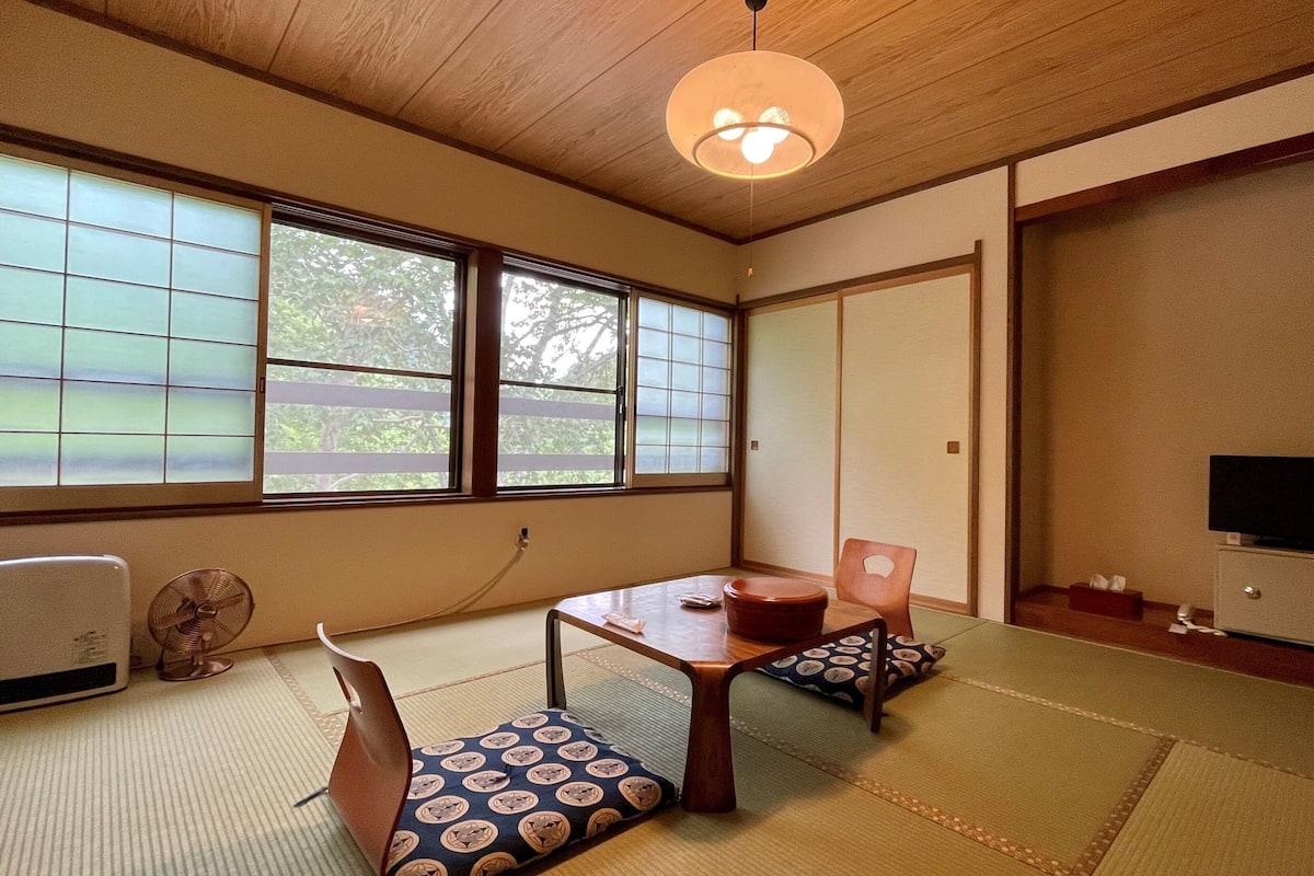 A traditional Japanese room with tatami flooring, a low table, a sofa, a fan, and a television.