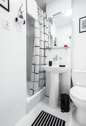 A modern bathroom with a white tiled floor, a black and white patterned shower curtain, a white sink, a toilet, and a black trash bin. - Elegant Apartment Panoramic (Warsaw)