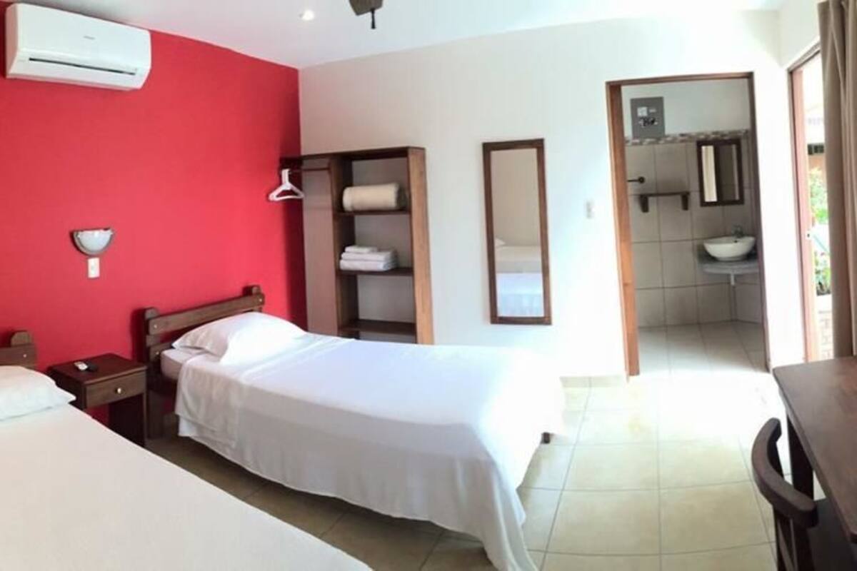 A hotel room with two beds, a red accent wall, a wooden nightstand, a desk, and a bathroom visible through an open door.