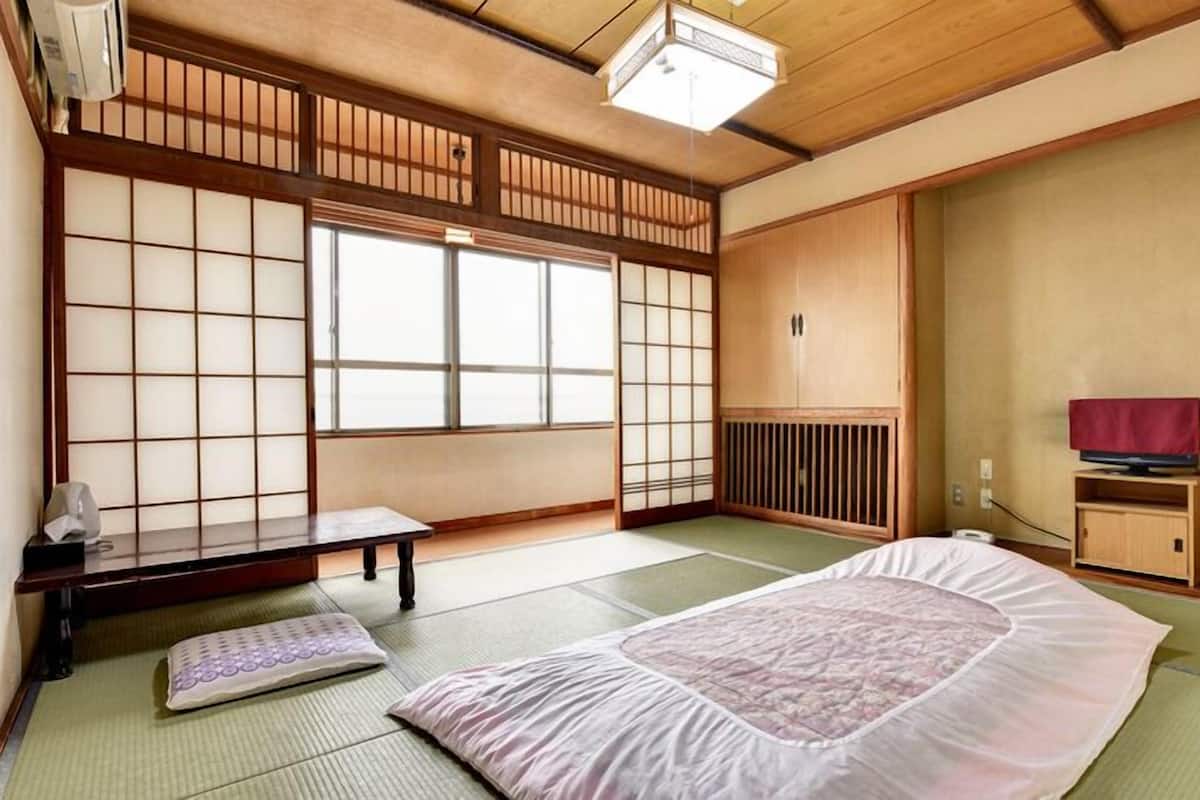 A traditional Japanese room with tatami flooring, a futon bed, a low wooden cabinet, a television, and a sliding shoji screen.
