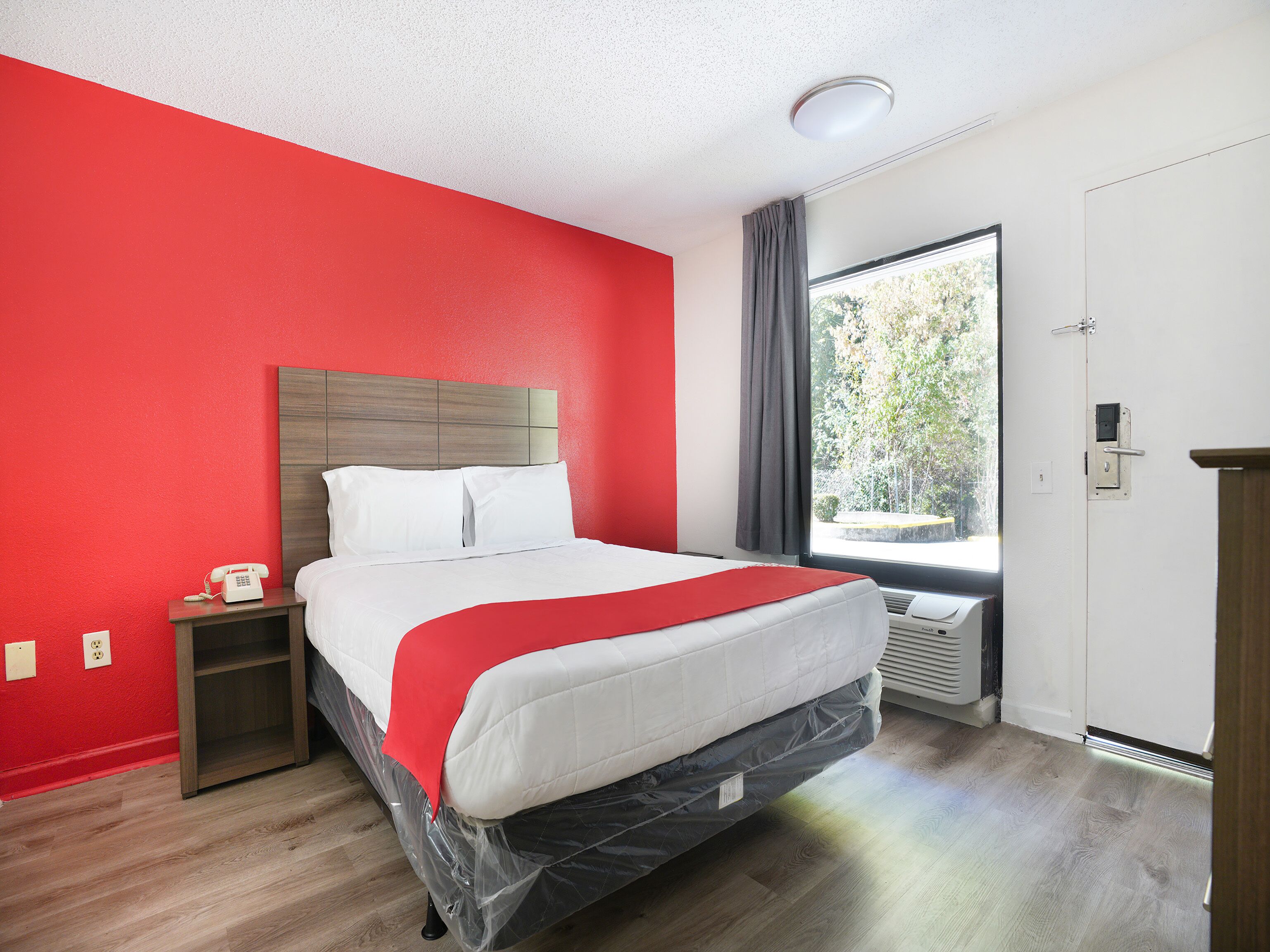 A hotel room with a bed, a nightstand, a telephone, a window with a view of trees, and a door with a lock.