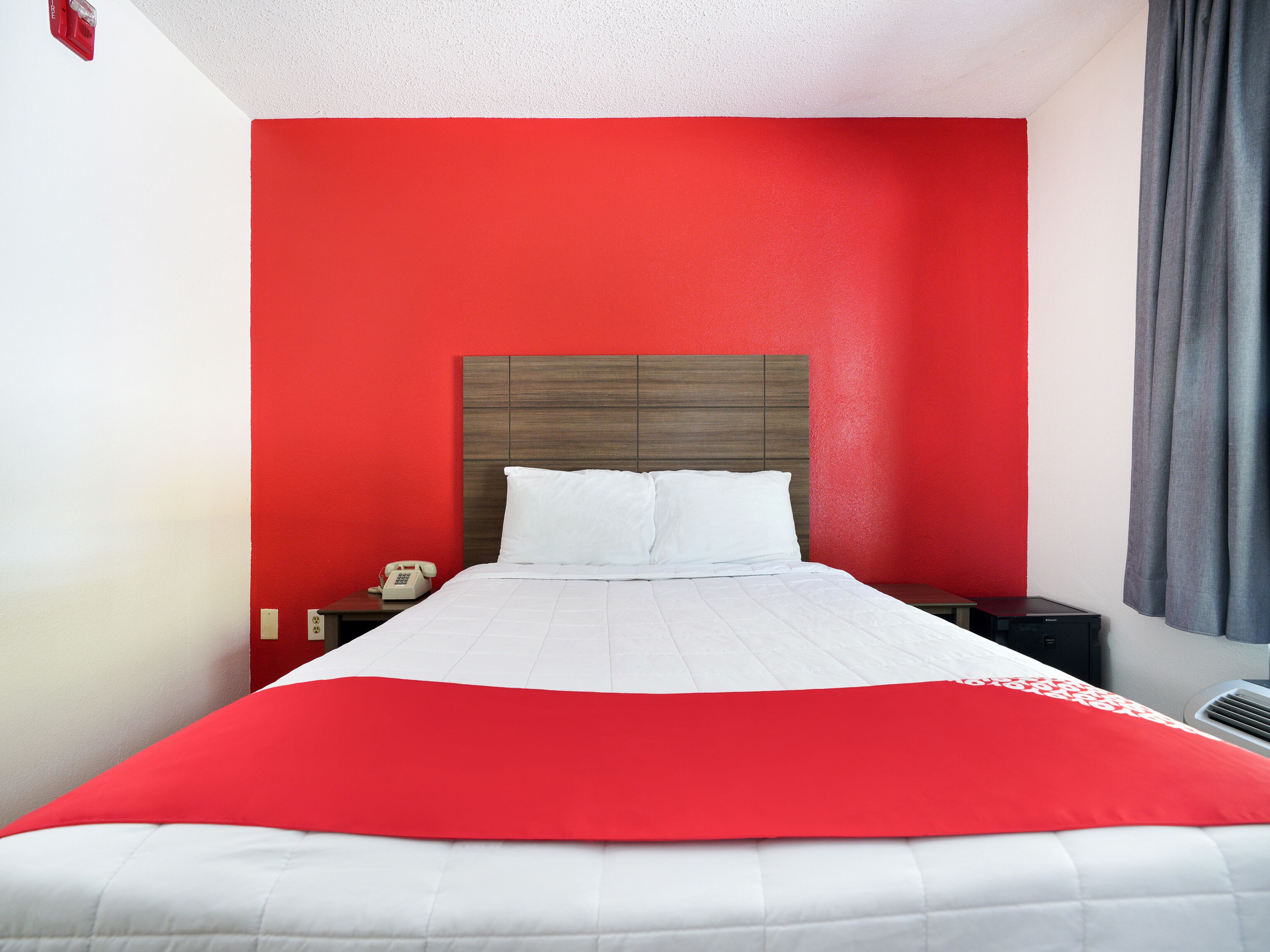 A hotel room with a red accent wall, a white bedspread, and a grey curtain.
