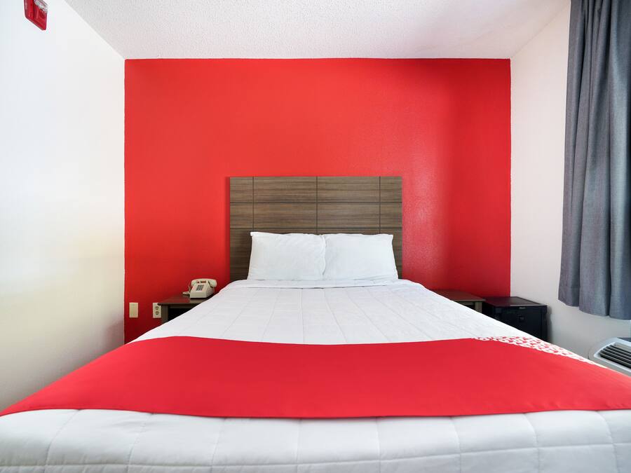 A hotel room with a red accent wall, a white bedspread, and a grey curtain.