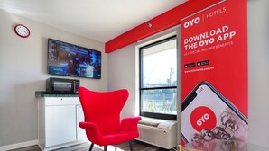 Una habitació d'hotel moderna amb una paret vermella d'accent, un gran finestral, una cadira vermella, un televisor de pantalla plana i un cartell promocional de l'aplicació OYO.