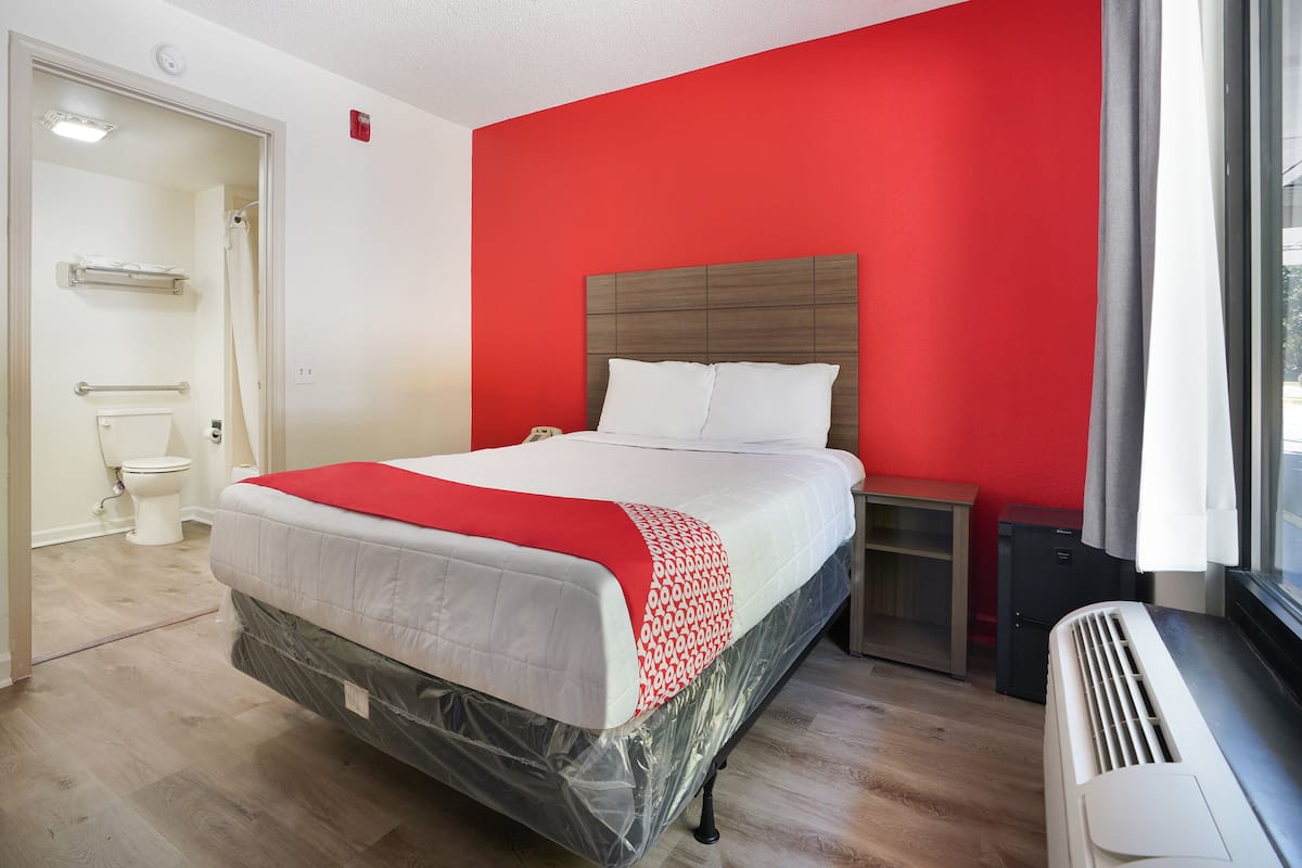 A hotel room with a bed, a bedside table, a heater, and a window with curtains.