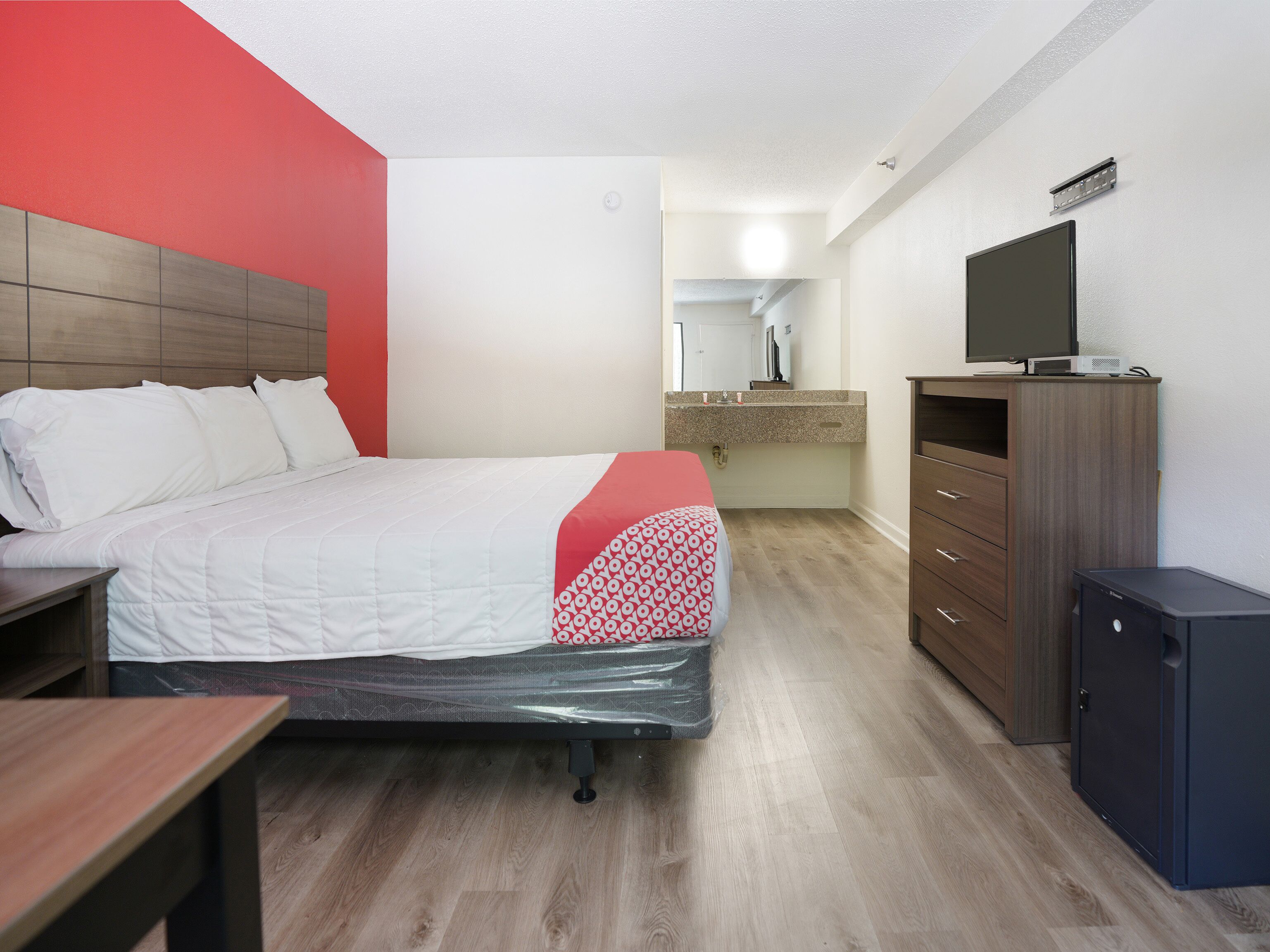 A hotel room with a bed, a desk, a dresser, a television, and a bathroom visible in the background.