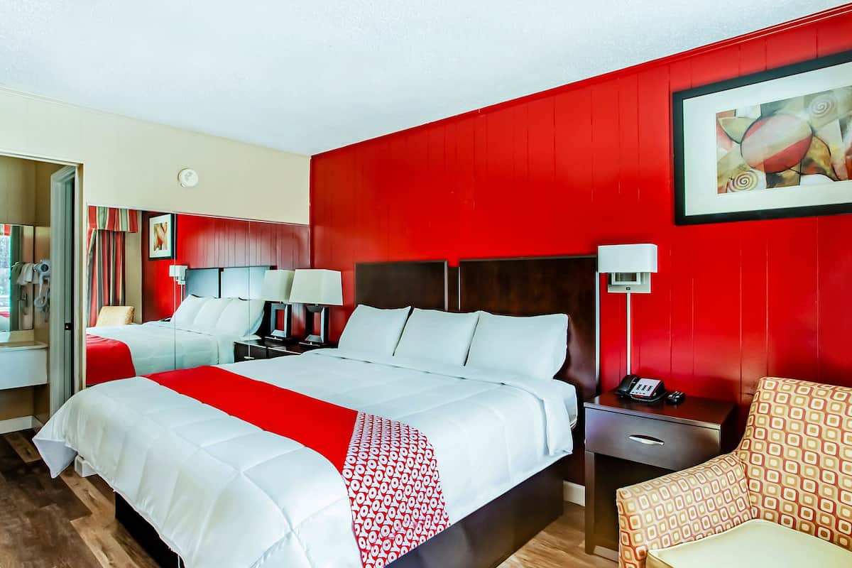 A hotel room with two beds, red walls, a painting, a telephone, and a chair.