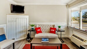 A modern hotel room with a sofa bed, a flat-screen TV mounted on the wall, a small table with a book, and a window with a view of the outside. - OYO Hotel Ridgeland East (Ridgeland)