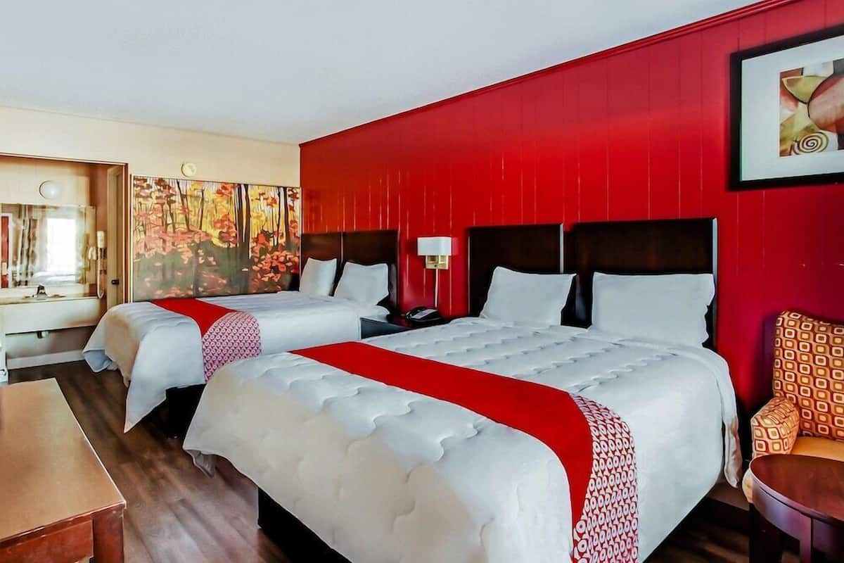 A hotel room with two beds, a red accent wall, a painting, and a mirror.