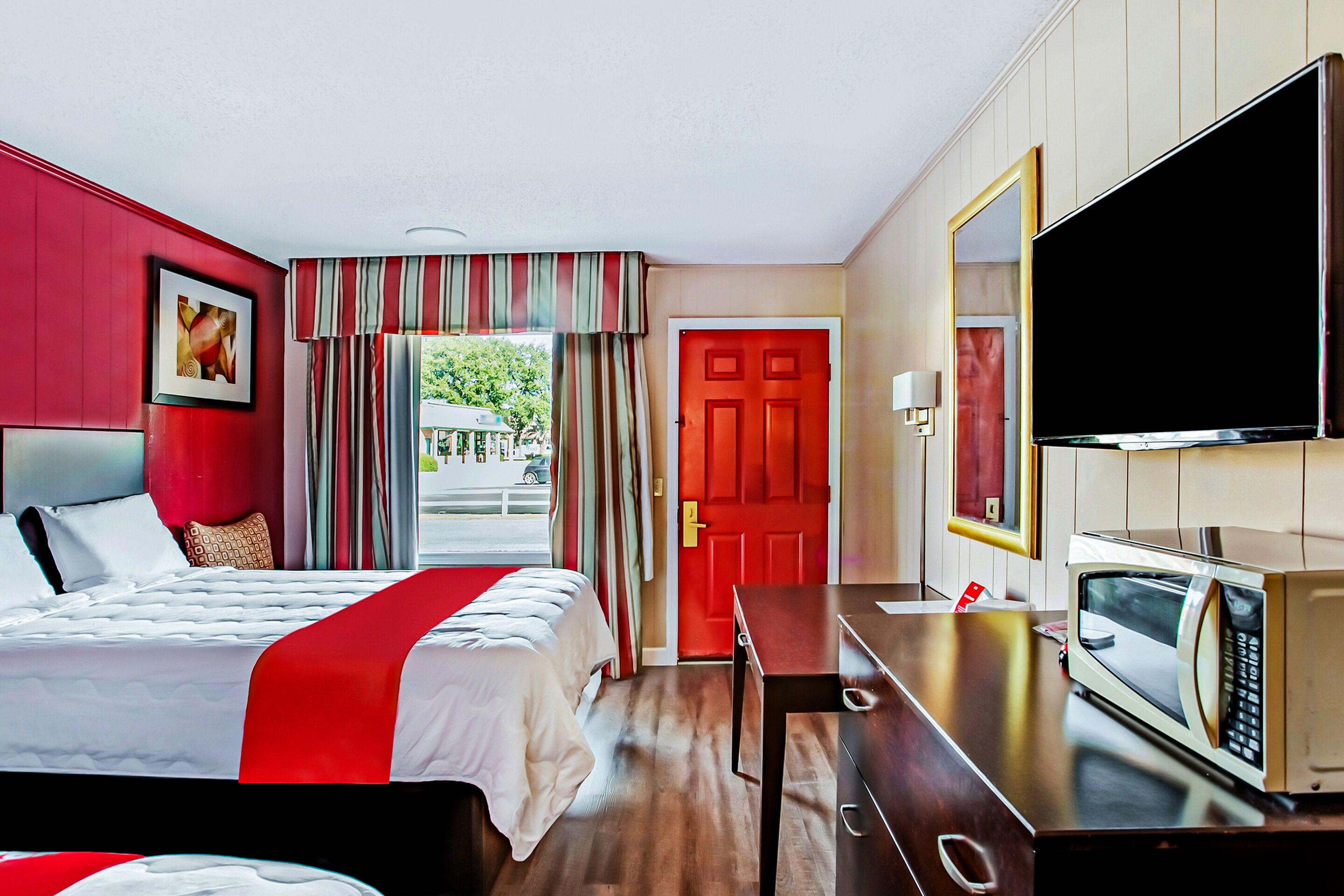 A hotel room with a bed, a desk with a microwave, a TV, and a red door.