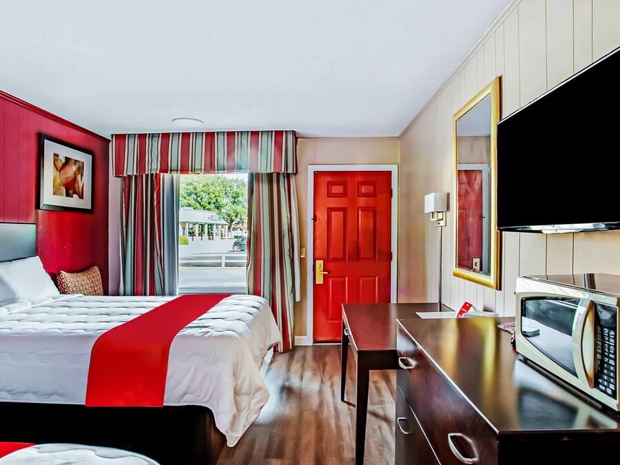 Ett hotellrum med en säng, ett skrivbord med en mikrovågsugn, en tv och en röd dörr.