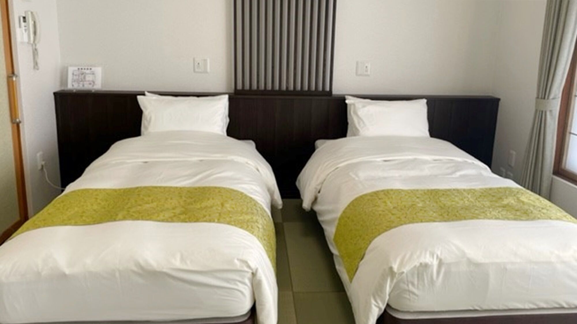 A hotel room with two beds, a window with curtains, and a wall-mounted air conditioning unit.